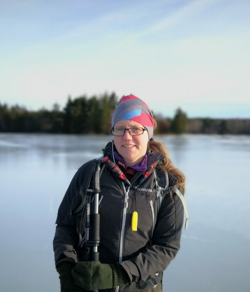 Syntolkning: En porträtt bild på Hanna. Blurrig bakgrund med frusen sjö och granskog. Hanna har isdubbar och ispik med sig och kisar lite mot solen. 